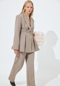 Blazer beige a medida con solapas de pico, doble botonadura y diseño plisado, combinado con pantalones a juego y un bolso rosa esponjoso.