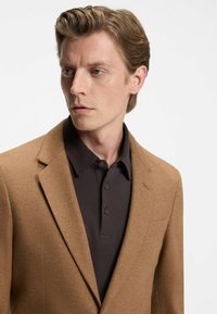 Blazer marrone in lana con rever classico, abbinato a una polo scura con colletto e bottoni. Tessuto liscio.