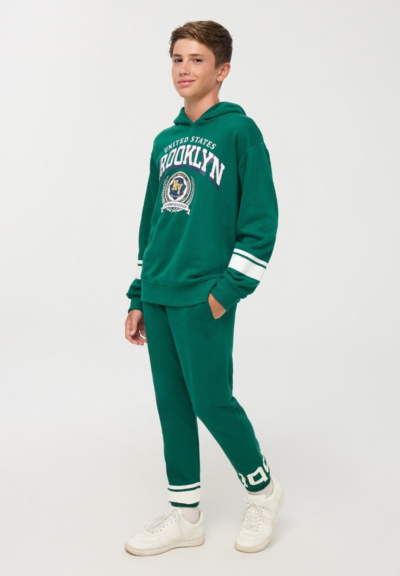 Felpa verde con strisce bianche e grafica di Brooklyn, abbinata a pantaloni verdi coordinati. Sneakers bianche completano l'outfit.