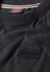 Tmavě šedá látka s vyšitým logem "Superdry" a částečně viditelnou modrou a oranžovou štítkovou nálepkou blízko límce.