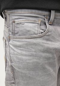 Vaqueros de mezclilla gris con un aspecto ligeramente desvaído, que presentan cinco bolsillos y detalles en costura naranja. La tela tiene un acabado suave y texturizado.