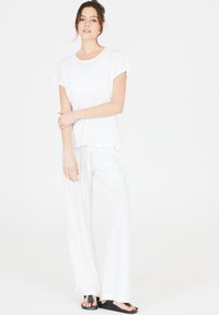 Valkoinen lyhythihainen T-paita pehmeästä kankaasta, yhdistettynä löysiin valkoisiin pellavahousuihin ja mustiin liukuvarvastossuihin, joissa on rento istuvuus.