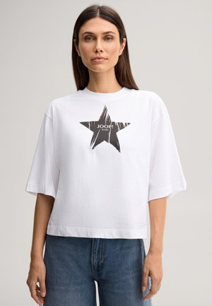 Vrouw draagt een wit oversized T-shirt met een zwarte ster en tekst "JOOP! JEANS", gecombineerd met blauwe spijkerbroek.