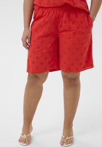 Femme portant un short rouge brodé et un haut assorti, debout les mains dans les poches, sandales beige avec détail coquillage doré.