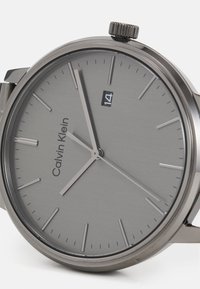 Grau gefärbte Edelstahl-Uhr mit minimalistischem Design, ausgestattet mit einem glatten Zifferblatt, dünnen Stundenmarkierungen und einer Datumsanzeige bei 14. Inklusive einer strukturierten Krone.