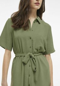 Aliejinės žalios spalvos marškinėlių suknelė trumpomis rankovėmis, su mygtukais per visą priekį, kišene ir diržu, kuriuo galima surišti liemenį, pasižymi tekstūruotu audiniu.