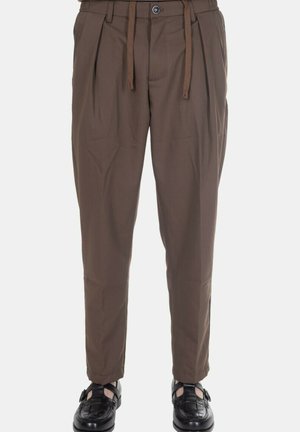 Pantaloni marroni plissettati con bottone e coulisse in vita, indossati con scarpe nere in pelle intrecciata con fibbia su sfondo bianco.