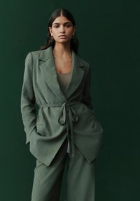 Olivgrüner Blazer aus strukturiertem Stoff, mit breiten Revers, gebundenem Gürtel und vorderen Taschen, getragen über einem figurbetonten Oberteil und passenden Hosen.