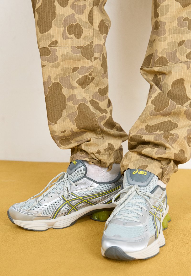 Baskets de sport gris clair avec des accents argentés, des touches de rembourrage vert et un dessus en maille texturée, associées à un pantalon à motif camouflage beige.