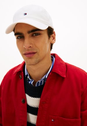 Bela baseball kapa z majhnim logotipom, rdeča jakna, modro-bela črtasta majica in mornarsko modra jopica z belimi črtami. Čista podlaga.