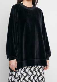 Felpa in velluto nero con maniche lunghe, scollatura tonda, spalle basse e una vestibilità comoda, abbinata a una gonna fantasia sottostante.