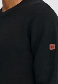 Pulover negru din tricot cu guler rotund, material texturat și o etichetă din piele maro pe mâneca stângă.