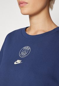 Mørkeblå sweatshirt med rund hals, brodert hvit Paris Saint-Germain-logo og hvit Nike swoosh nær kanten.