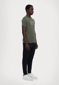 Grønn kortermet t-skjorte med gull EA7-logo, kombinert med mørk navy joggebukser og hvite sneakers. T-skjorten er tettsittende og laget av bomull.
