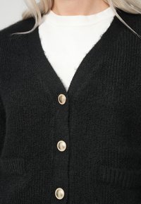 Juodas mezgtas kardiganas su V formos kaklu, pasižymintis tekstūruotu audiniu ir aukso spalvos sagomis. Įtrauktos dvi priekinės kišenės, kad suteiktų papildomą detalių.