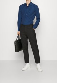 Mörkblå skjorta med knappar och subtilt mönster, i kombination med svarta skräddarsydda byxor och vita sneakers. En svart väska hålls i vänster hand.