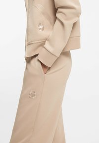 Beżowa kurtka na zamek o luźnym kroju, z haftowanymi akcentami logo oraz bocznymi kieszeniami w dopasowanych spodniach. Gładka tekstura materiału.