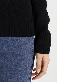 Hand ruht neben blauen Jeans aus Denim, trägt einen schwarzen, gerippten Pullover mit langen Ärmeln.