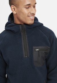 Marinefleece pullover med hætte, der har en halv lynlås foran og en kontrasterende sort lomme med lynlås lukning på brystet. Blød tekstur.