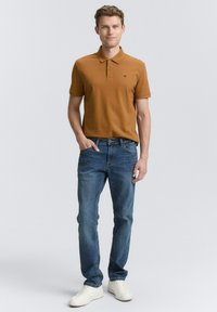 Mand med kort hår iført en brun polo-shirt, blå jeans og hvide sneakers står foran en lys grå baggrund.