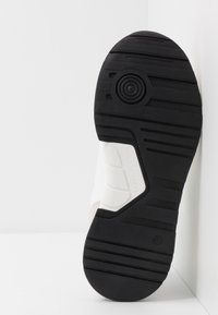 Semelle extérieure en caoutchouc noir avec motif de bande de roulement texturé, semelle intermédiaire blanche et détail de marque. Profil latéral montrant la forme et les caractéristiques de construction.