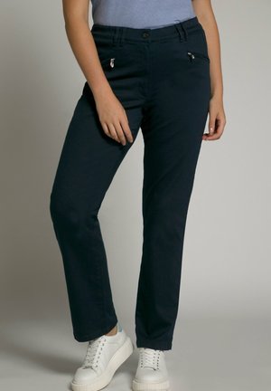 Kvinde iført mørkeblå bukser med lige ben og lynlåslommer foran, lyseblå top og hvide platformsneakers mod en ensfarvet baggrund.