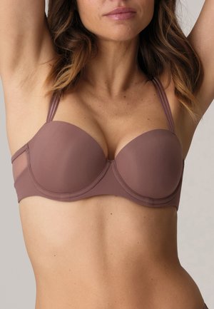Donna che indossa un reggiseno senza spalline color malva con le braccia sollevate, mostrando la parte superiore del torso e la texture naturale della pelle su uno sfondo neutro.