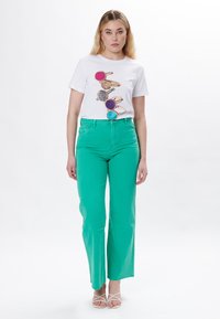 Camiseta blanca con un gráfico de formas redondas coloridas y brillantes; combinada con jeans de pierna ancha de cintura alta en un brillante color verde y sandalias blancas con tiras.