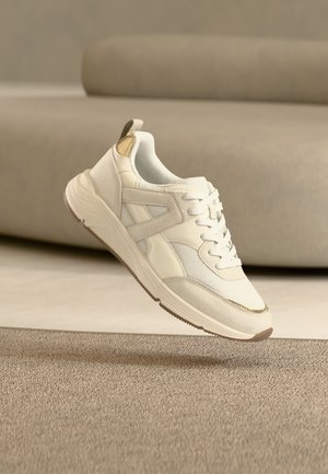 Λευκό και μπεζ αθλητικό παπούτσι με λεπτομέρειες από δίχτυ και σουέτ, αιωρούμενο πάνω από μπεζ υφασμάτινο χαλί σε δωμάτιο με ουδέτερους τόνους.