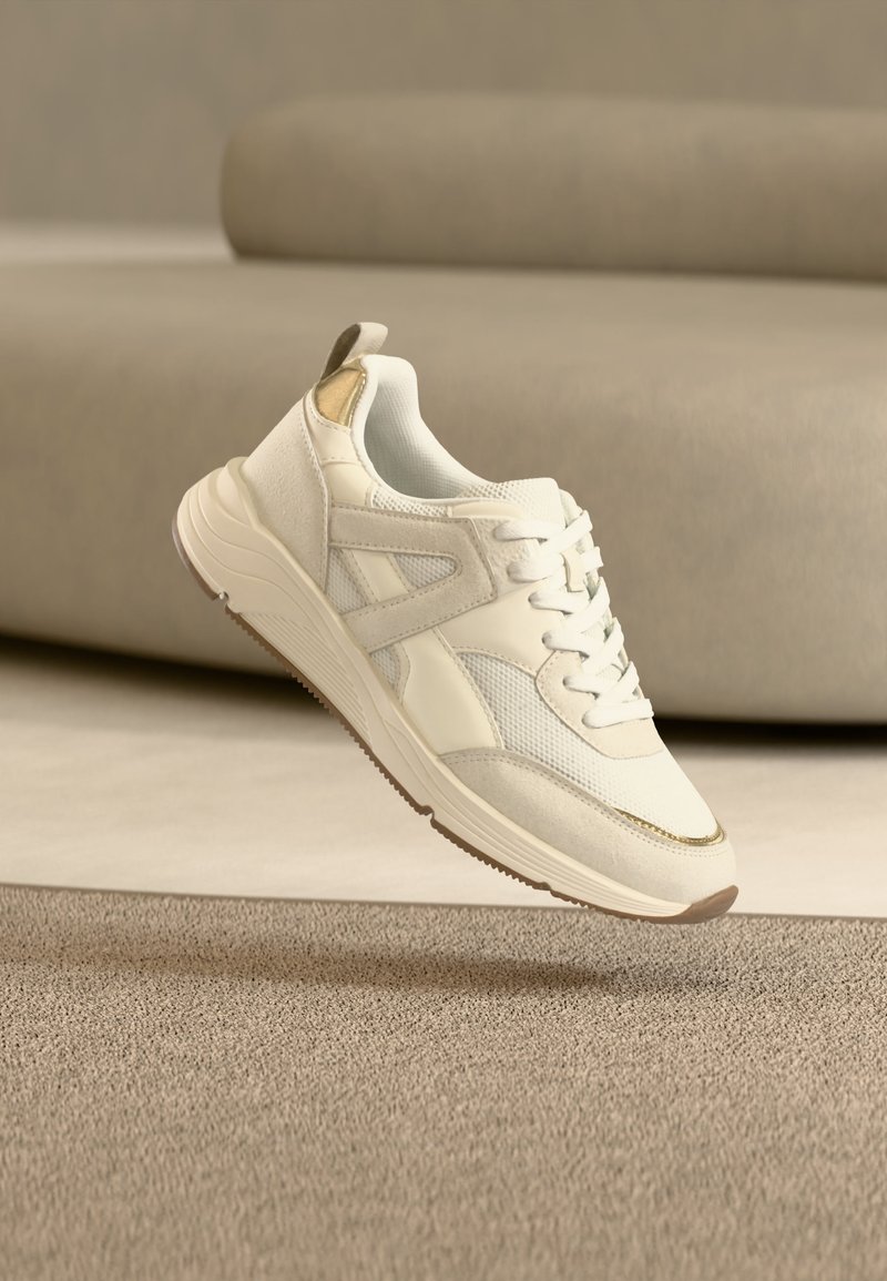 Λευκό και μπεζ αθλητικό παπούτσι με λεπτομέρειες από δίχτυ και σουέτ, αιωρούμενο πάνω από μπεζ υφασμάτινο χαλί σε δωμάτιο με ουδέτερους τόνους.