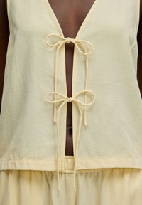 Lichte gele mouwloze top met een diepe V-hals, voorzien van twee strikken bij de halslijn en een geplooide zoom. Zachte, gestructureerde stof.