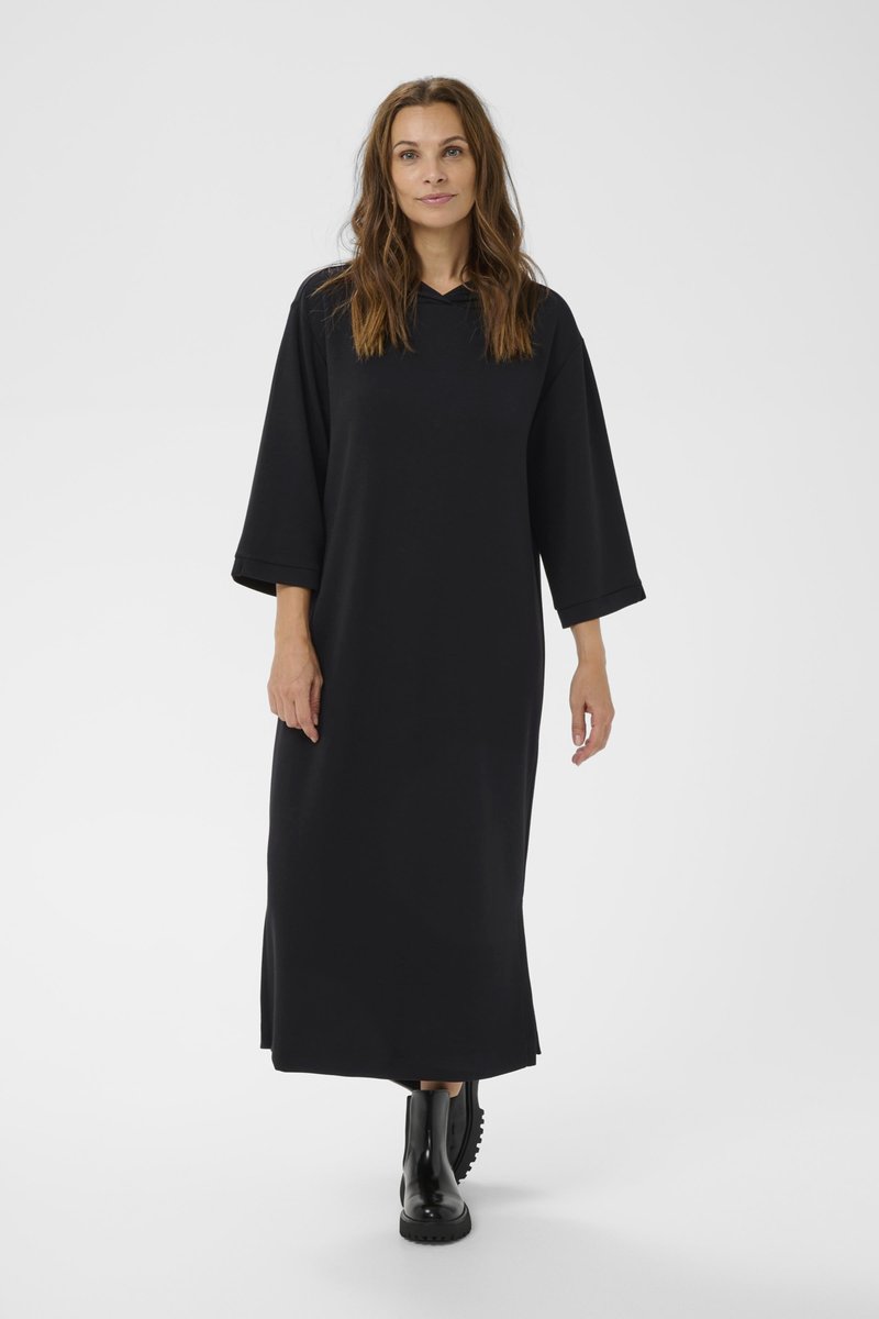 Robe maxi noire avec de longues manches larges et une capuche, présentant une coupe décontractée. Portée avec des bottes noires à talons épais pour un contraste.