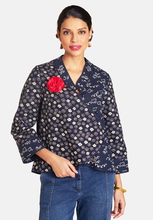 Frau mit dunklen Haaren trägt eine marineblaue, geblümte Wickelbluse mit einer roten Stoffblumen-Brosche und blauen Jeans, posiert mit einer Hand in der Tasche.