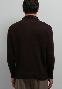 Man met een donkergroene gebreide trui met een geribbelde kraag, manchetten en zoom, die met zijn rug naar een lichtgrijze muur staat gekeerd.