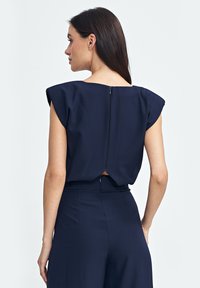 Granatowa bluzka ma krótkie rękawy, niski tył z widocznym zamkiem błyskawicznym oraz luźny krój. Materiał ma gładką fakturę.