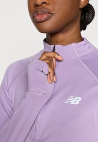 Αθλητική μπλούζα ανοιχτόμωβ με φερμουάρ στο ένα τέταρτο του γιακά, με μανίκια με οπή για τον αντίχειρα και λευκό λογότυπο New Balance στο στήθος.