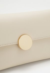 Pochette en cuir beige à texture lisse, dotée d'un fermoir circulaire doré proéminent et de détails de couture subtils.