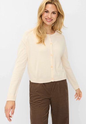 Smilende kvinde med langt blond hår iført en cremefarvet cardigan med knapper og brune fløjlsbukser, stående mod en ensfarvet lys baggrund.