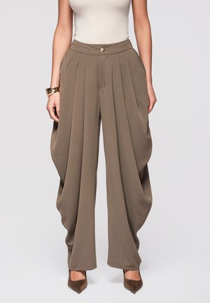 Donna che indossa pantaloni taupe a vita alta, ampi con pieghe e lati drappeggiati, abbinati a tacchi appuntiti marroni e una canottiera bianca senza maniche.