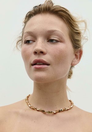 Collier doré avec des formes irrégulières texturées et des accents verts, présentant des surfaces lisses et réfléchissantes. Design courbé, porté près du cou.