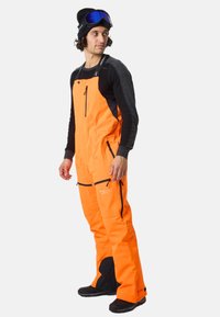 Felle oranje sneeuwoverall met zwarte accenten, ritszakken en verstelbare schouderbanden. Gemaakt van duurzame materialen voor gebruik in koude weersomstandigheden.