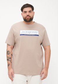Beige bomulls t-skjorte med korte ermer, med en blå og hvit grafikk med teksten "JACK & JONES" på forsiden. Glatt tekstur.