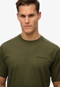 Oliven grøn kortærmet t-shirt lavet af bomuld med rund hals, med "SUPERDRY" logoet trykt i sort på fronten.