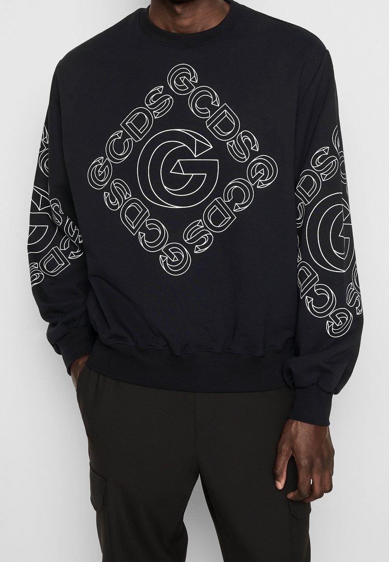 Sweat-shirt noir avec logos "GCDS" en contour blanc disposés en forme de losange sur le devant et répétés sur les manches, porté avec un pantalon noir.