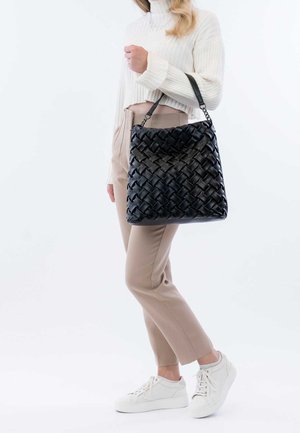 Femme portant un pantalon beige et un pull blanc, tenant un grand sac à main en cuir tressé noir sur l'épaule, chaussée de baskets blanches.