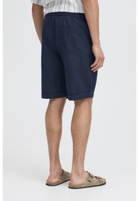 Mørkeblå linnedshorts med elastisk talje, opkrogede kanter og en enkelt baglomme. Båret med lysebrune sandaler.