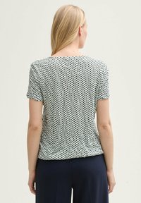 T-shirt à manches courtes en tissu vert clair avec un motif à pois noirs, encolure ras du cou et coupe décontractée, associé à un pantalon foncé.