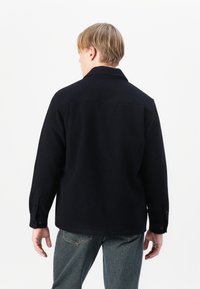 Navy wool shirt jacket with a slightly textured finish, collared neck, and buttoned cuffs, worn over faded denim jeans.