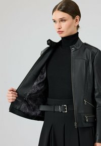 Zwarte leren jas met zilverkleurige accenten, voorzien van een patroonvoering, ritszakken en een getailleerd ontwerp. Draagt over een zwarte coltrui.