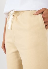 Pantalons de survêtement beige en tissu doux avec une taille élastique, dotés d'un cordon de serrage et de poches latérales, portés avec un haut blanc.